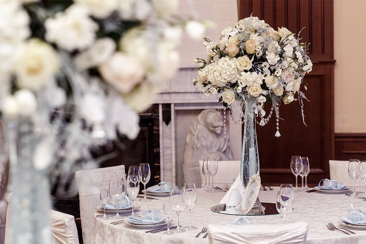 Salón de bodas elegante con mesas decoradas, centro floral blanco en altura y detalles en cristal.